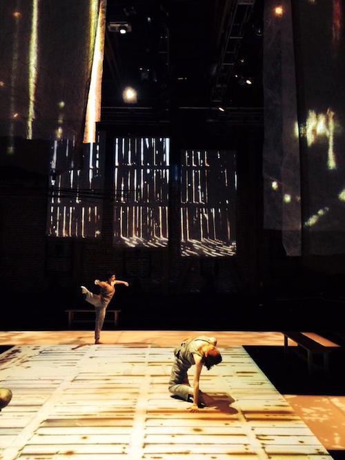 Two dancers, one on their knee and the other balancing on one leg in front of the projections. The floor also has a projection of wood slates